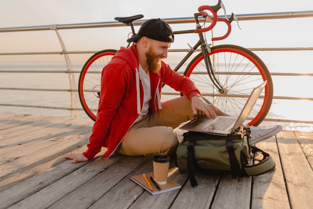 Homem sentado em um deck de madeira, com um notebook apoiado em cima de sua mochila. Logo atrás dele, há uma bicicleta, encostada em uma cerca. Ao fundo, vemos o mar.