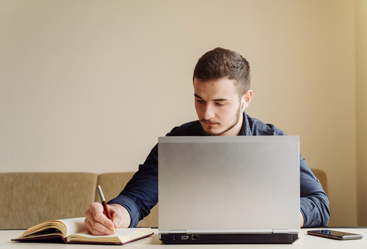 Homem sentado usando um laptop. Ele está anotando algo em um caderno, enquanto olha para baixo. Parece estar trabalhando ou estudando.