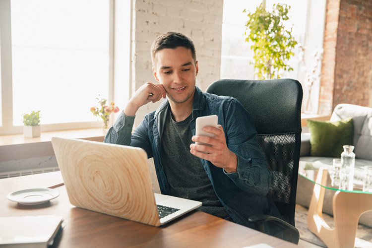 Homem sentado em uma cadeira de escritório, segurando um smartphone com uma mão. A sua frente, uma mesa com notebook. O homem olha em direção ao smartphone e sorri.