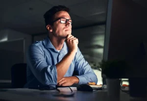 Homem observando o monitor de um computador. Ele está com uma mão no queixo, demonstrando pensar em algo. Ele usa óculos e camisa social.